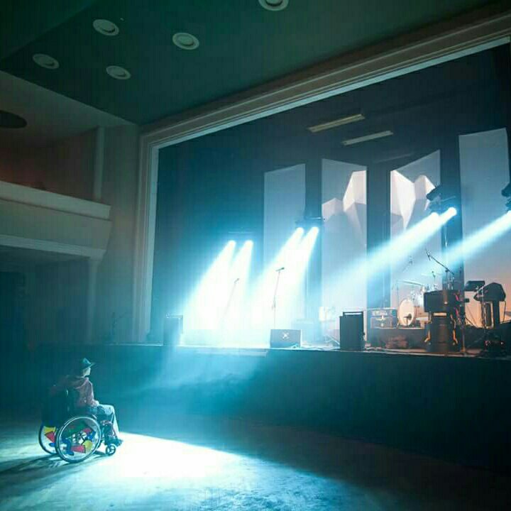 November_Crash steht im Rollstuhl in einer großen Halle und schaut auf eine Bühne. Von der Bühne leuchten Scheinwerfer auf sie. Auf der Bühne stehen verschiedene Musikinstrumente.   
