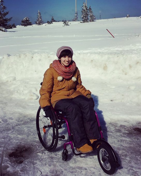 Lisa sitzt im Rollstuhl. Um sie herum ist Schnee. Sie trägt Mütze, Jacke, Hose,Schuhe, Schal und Handschuhe in schwarz und Orangentönen. Vorne am Rollstuhl ist ein 5. extra Rad montiert. 